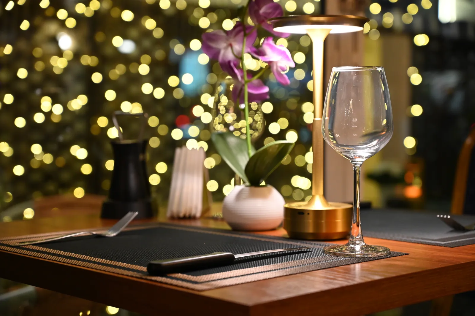 Table de restaurant dressée le soir, verre à vin et petite lampe, lumières en arrière-plan